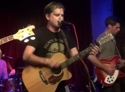 White man with brown hair playing a guitar and singing at a mic with other musicians