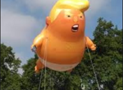 Big inflated balloon shaped like a fat baby in a diaper with orange skin and large yellow hair flowing at the top with his mouth open held up high in the air by strings