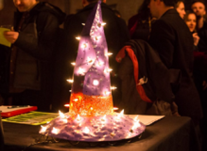 People at a party in the background and on a table in front a sparkly purple pointy hat with a wide brim and with an orange band around it