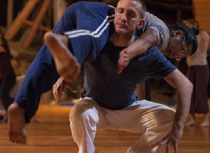 White middle aged man squatting with his eyes closed holding a woman across is back in a dance move