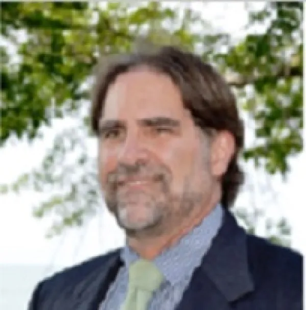 Middle aged white man with short brown hair, a grayish beard and mustache wearing a blue suit standing in front of a tree