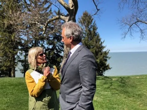 Short blonde wtoman wearing a yellow coat and sunglasses talking and gesturing to a taller white man with gray hair and a gray suit outside by tree and a lake 