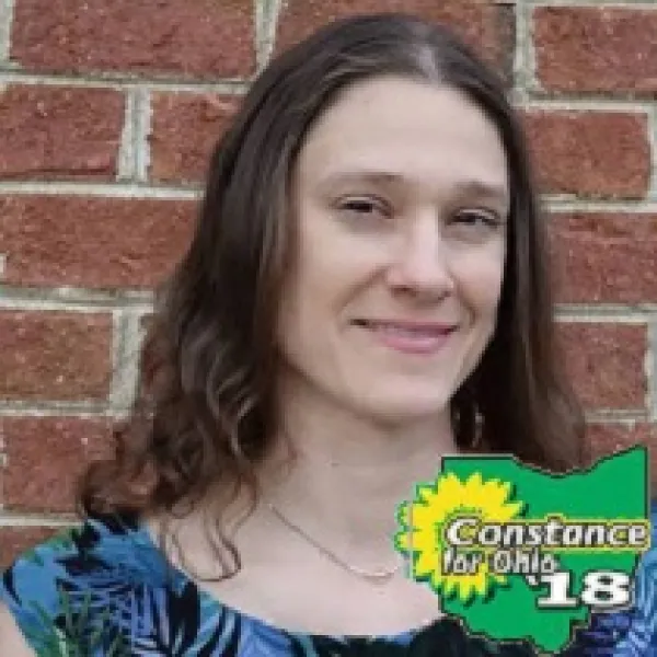 Young white woman with brown curly hair smiling standing in front of a brick wall with the words Constance for Ohio '18 in the right side bottom