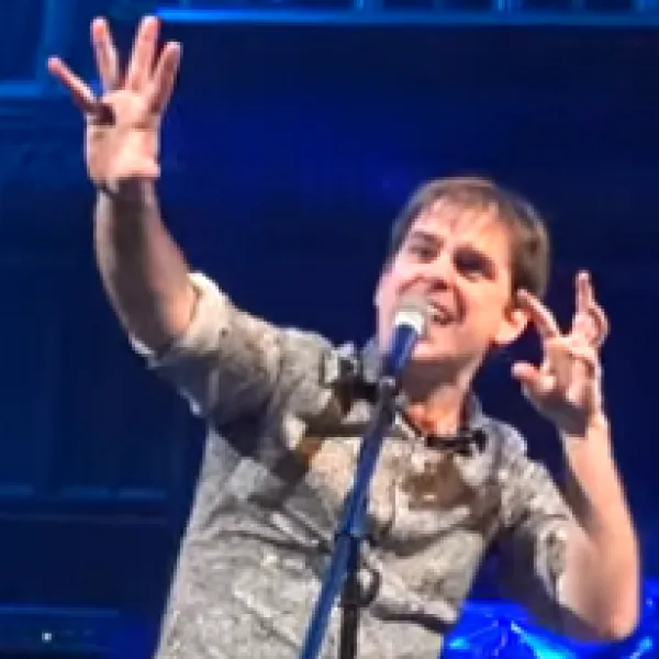 Young white man with both arms up in the air singing at a mic