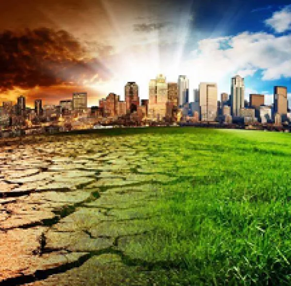 A cityscape against a horizon with sunrays shooting from the ground to a blue sky on right and fiery sky on left with the fore ground at right green grass and at left cracked and dry