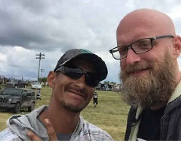 Bearded white man with glasses an bald head smiling with a dark-skinned man in sunglasses and a baseball cap outside in front of some cars and a cloudy sky