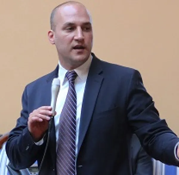 Baldish white man wearing a suit holding a microphone and talking