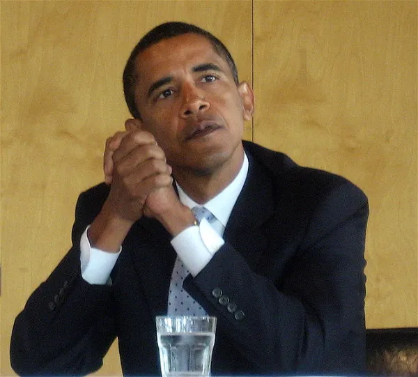 Obama with elbows on table clasping his hands together looking pensive