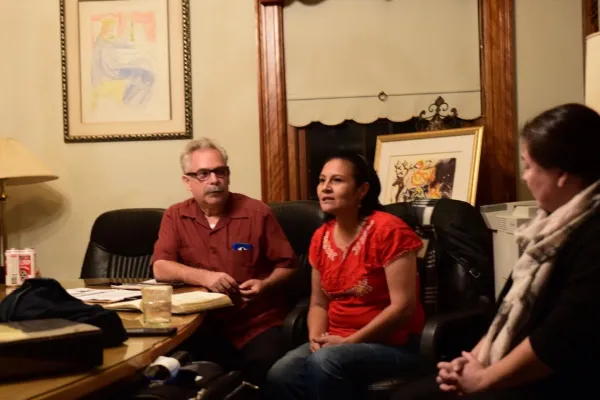 Gray haired man with black-rimmed glasses sitting at a table looking at a dark haired woman who is speaking also sitting in a chair