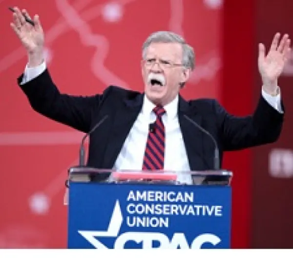 Older man with gray hair and long white mustache standing at a podium with his mouth  open waving both hands in the air and the words American Conservative Union on the podium