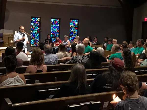 Man in white shirt talking into a mic at the far left side and a congregation of people sitting in pews and three stained glass windows in the back