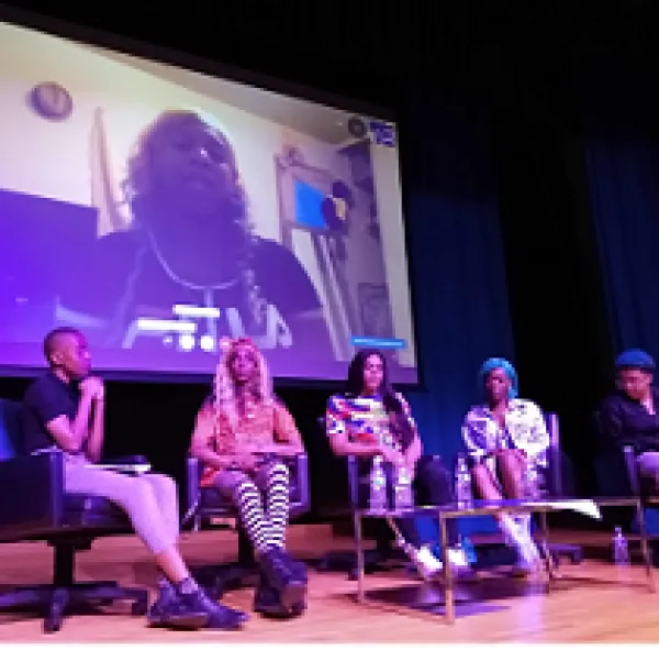 Stage with large screen with a black trans woman speaking and five black women in chairs below