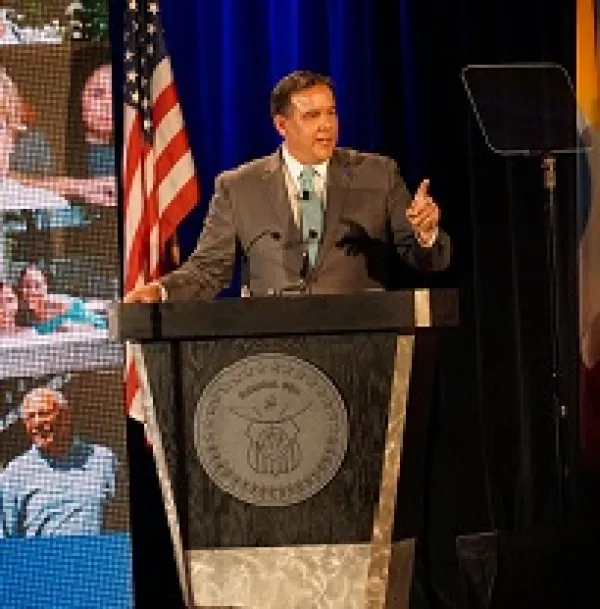 White middle-aged man in a brown suit at a podium pointed and speaking with an U.S. flag and a screen with pictures behind him