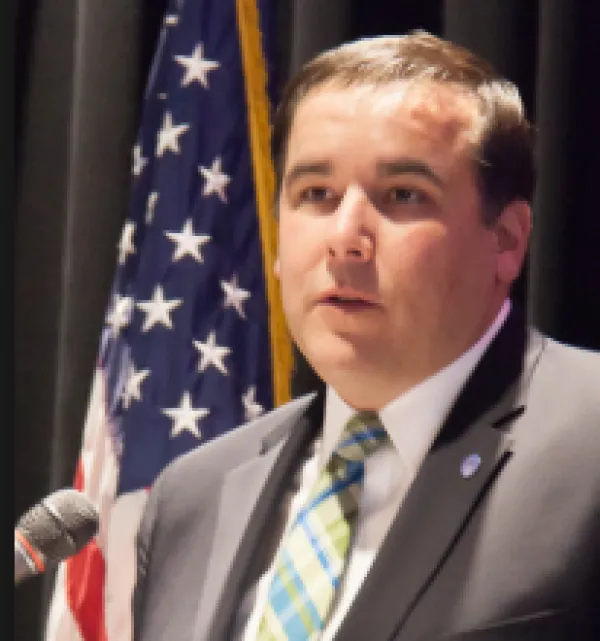 Middle aged white man in a suit with brown receding hair at a mic with a flag in the background