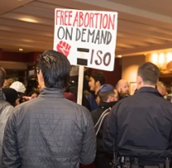 Backs of two men, one with a cop looking uniform on and the other with a sign that says Free Abortion on Deand ISO with a red fist
