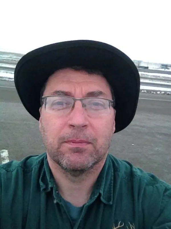 Man with glasses and wide brimmed black hat, looking grim