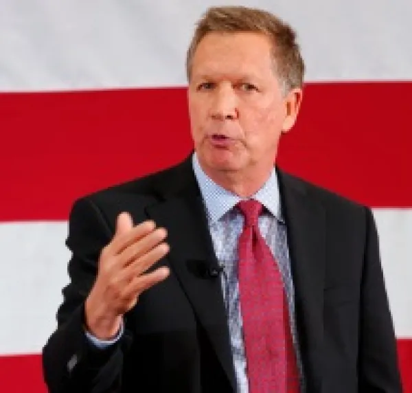 White man middle-aged in dark suit with red tie and white shirt talking and holding up his hand by his chest in front of a red and white striped background