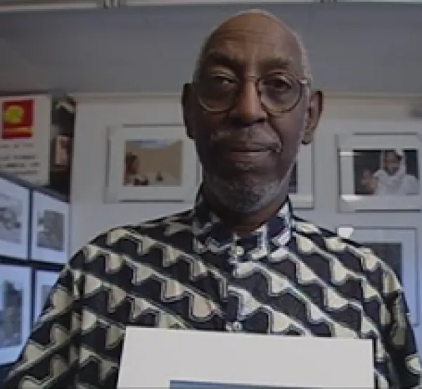 Older black man, bald with gray mustache and beard in a gray and white shirt holding a photo and standing in front of lots of other photos on the wall. 
