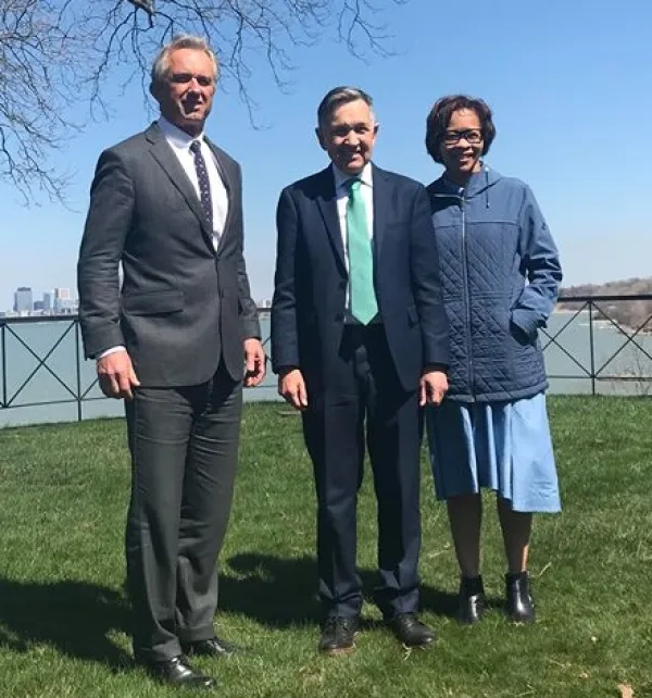 Three people standing outside on the grass with  body of water behind them, a tall white man in a suit with gray hair, and shorter white man in a suit and a black woman with glasses
