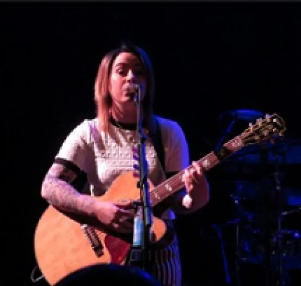Young white woman in a white t-shirt playing a guitar and singing at a mic with a black background