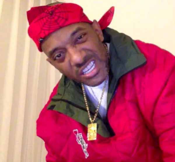 Black man in red jacket with red doo rag scarf on head with gold necklace, making a sneering face