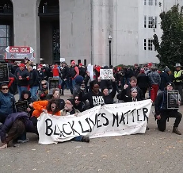 People wearing winter clothes bending down on one knee outside holding a long white banner with black letters reading Black Lives Matter in front of a large white building