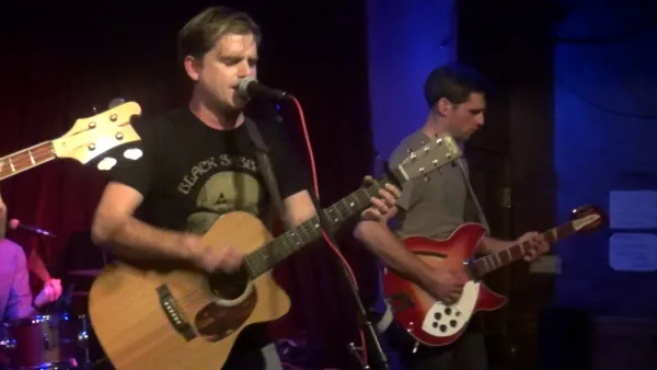 White man with brown hair playing a guitar and singing at a mic with other musicians
