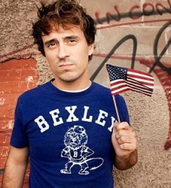Young  white man  with tousled brown hair wearing a blue T-shirt that says BEXLEY in white letters and he's waving a very tiny flag and kind of sneering at the camera in a bored way