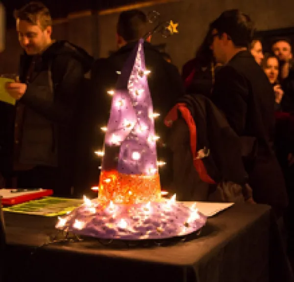 People at a party in the background and on a table in front a sparkly purple pointy hat with a wide brim and with an orange band around it