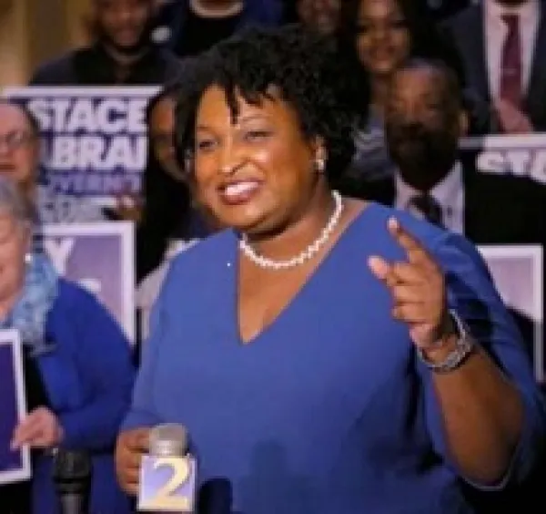 Older black woman with short curly black hair, white pearl necklace, blue dress, standing and smiling while gesturing in front of lots of people holding signs that say Stacey Abrams